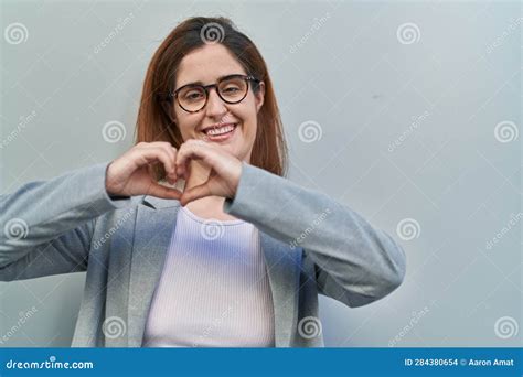 Brunette Woman Standing Over Grey Background Smiling in Love Doing Heart Symbol Shape with Hands ...