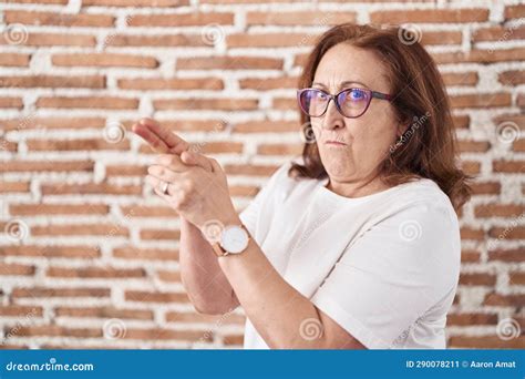 Senior Woman with Glasses Standing Over Bricks Wall Holding Symbolic Gun with Hand Gesture ...