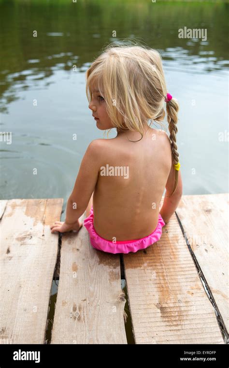 Rear view of child blond girl sitting on pier looking at view of pond ...