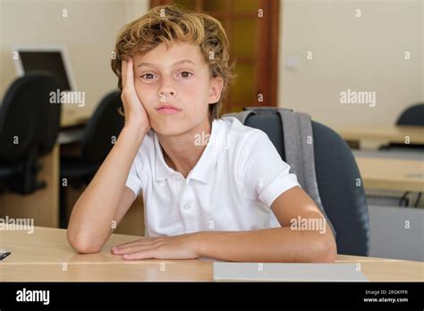 Bored schoolkid leaning on hand at wooden desk in classroom and looking at camera Stock Photo ...