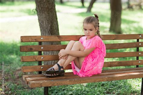 Little caucasian girl sitting in park on bench. A child in a beautiful pink dress in the open ...