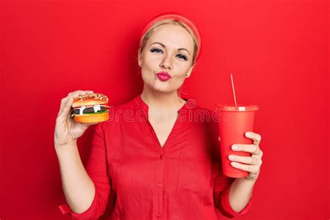 Young Blonde Woman Eating a Tasty Classic Burger with Fries and Soda Looking at the Camera ...