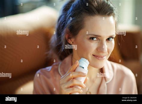 smiling young housewife in pajamas at modern home in sunny winter day using electric facial ...