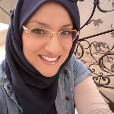 a woman wearing glasses and a hijab smiles at the camera while standing in front of a wrought ...