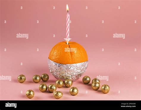 Fresh orange in aluminium foil with a lighted candle and with golden balls around, on pink ...