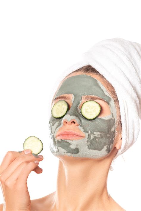 Beautiful young woman with clay mask on her face holding slices of fresh cucumber, skin care and ...