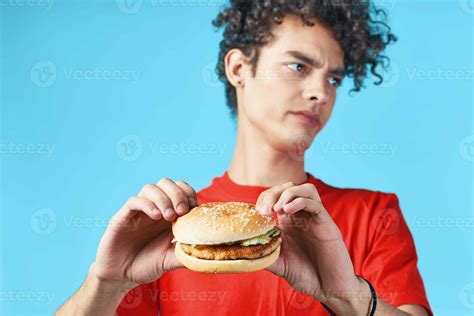 Cheerful guy with curly hair in a red t-shirt with a hamburger in his hands fast food diet ...