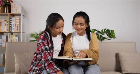 Happy asian twin girls reading a book and sitting on couch in living