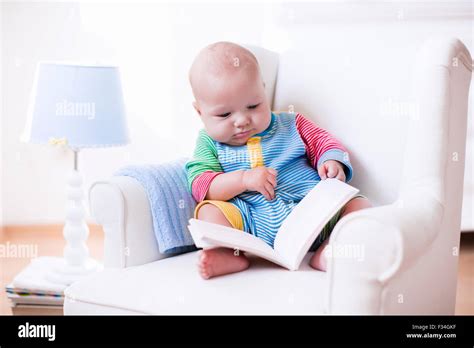 Cute funny baby boy reading a book sitting in a white chair at home ...