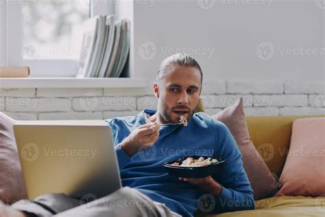 Handsome young man eating lunch and looking at laptop while sitting on