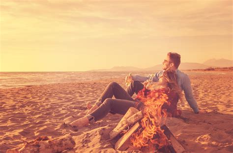 Young Couple Sitting On The Beach beside Campfire drinking beer