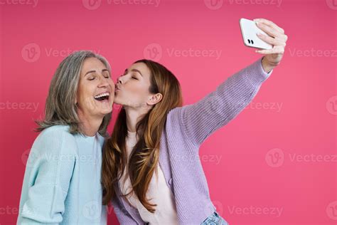 Adult daughter kissing her mother while making selfie against pink