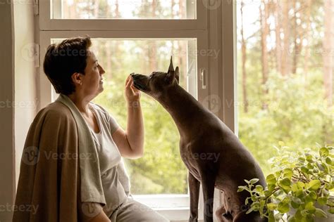 Middle age beautiful woman sitting on windowsill with her dog. 50-year